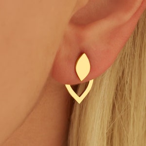 May include: Gold-colored earrings with a leaf-shaped stud and a V-shaped backing. The earrings are shown on an ear with blonde hair in the background. The earrings have a minimalist design.