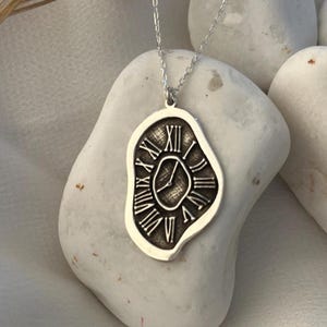 May include: A silver-toned pendant necklace featuring an irregular, melted-shape charm with a dark, recessed clock face design. The clock face displays Roman numerals and two hands, suspended from a delicate, thin silver-toned metal chain.