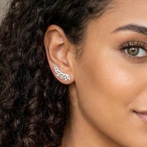 May include: A silver ear climber earring featuring a delicate, multi-pronged leaf or wing design that follows the curve of the earlobe. The metallic piece has a polished finish and is worn on the outer edge of the ear, creating a sleek look.