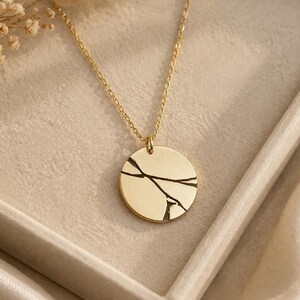May include: A gold-toned necklace featuring a circular pendant with a black engraved branch design. The pendant hangs from a delicate gold-toned chain. The jewelry piece is displayed on a light-colored surface with a small cluster of dried flowers.