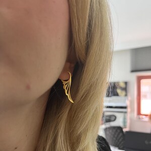 以下が含まれることがあります： 金色の羽の形をしたイヤリングが耳に装着されています。イヤリングは、複数の曲線と滑らかな仕上がりを備えた繊細でオープンなデザインです。イヤリングはスタイリッシュなアクセサリーです。