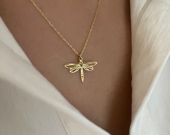 Puede incluir: Un collar de tono dorado con un delicado colgante de libélula geométrica con alas caladas y un cuerpo esbelto. El colgante cuelga de una cadena fina de tono dorado a juego que descansa sobre una camisa blanca de cuello.