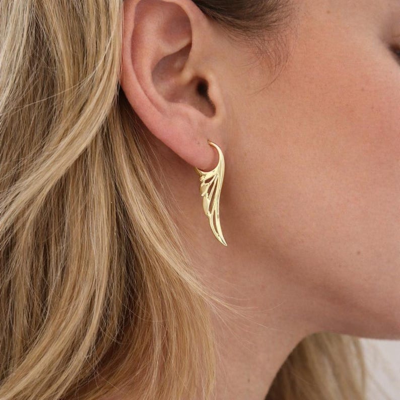 May include: A gold-colored earring in the shape of a wing is shown. The earring is attached to the ear. The wing design has detailed feather-like cutouts. The earring is a hoop style, with a curved design that follows the ear's shape.