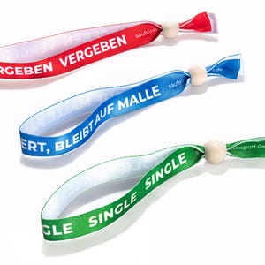 Puede incluir: Tres pulseras de colores con cuentas de madera. La pulsera roja dice "VERGEBEN", la pulsera azul dice "ERT, BLEIBT AUF MALLE" y la pulsera verde dice "GLE SINGLE".