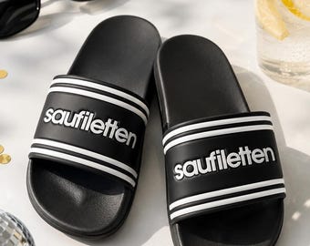 May include: A pair of black slide sandals featuring a wide strap with two horizontal white stripes and the word "saufiletten" embossed in white lettering. The sandals have a contoured footbed and a smooth, matte finish on the upper strap.