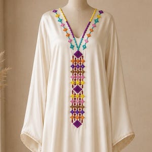 May include: A floor-length cream-coloured kaftan dress featuring long, wide sleeves and a V-neckline. The neckline and centre front are decorated with a vertical panel of intricate, colourful geometric embroidery in shades of purple, orange, yellow, and blue.