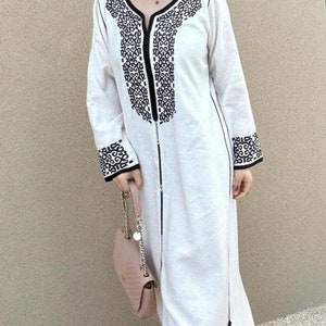 May include: A white long-sleeved dress with black embroidery around the neckline and cuffs. The dress has a zipper down the front and a small handbag is hanging from the wearer's arm.