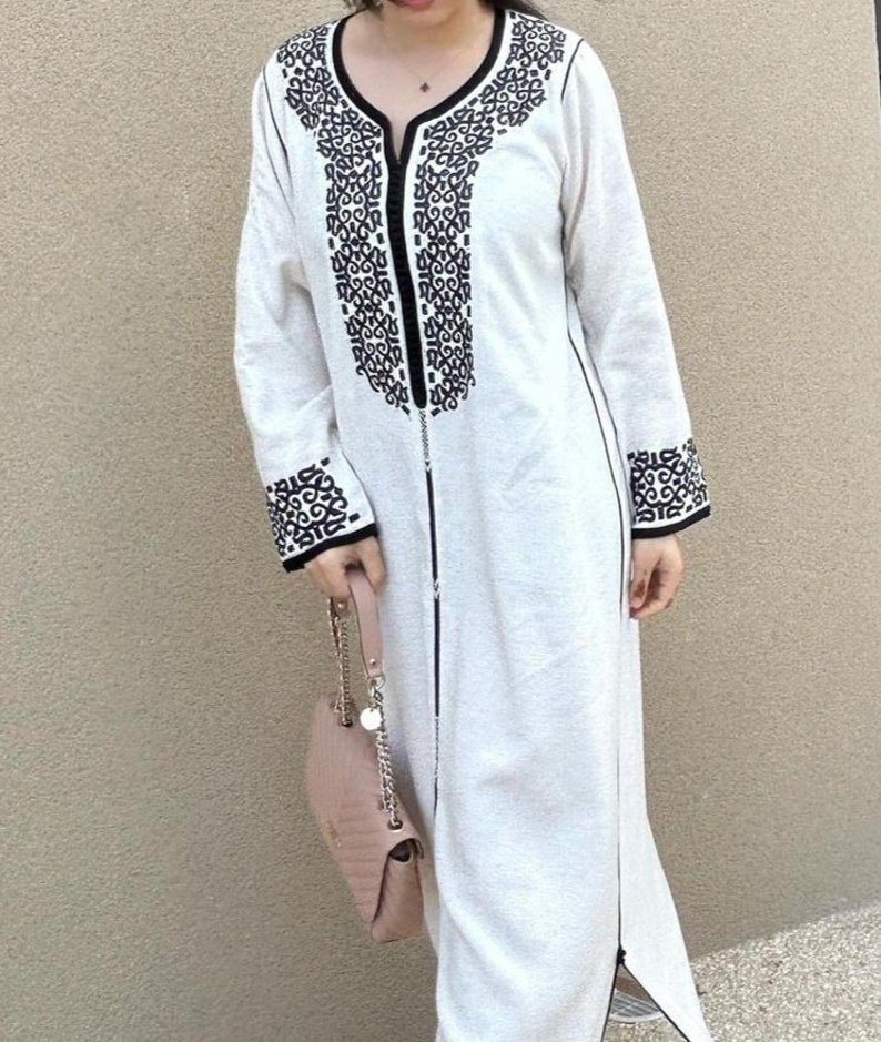 May include: A white long-sleeved dress with black embroidery around the neckline and cuffs. The dress has a zipper down the front and a small handbag is hanging from the wearer's arm.