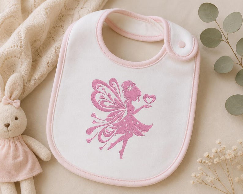 May include: A white baby bib with a soft pink trim and a snap closure. The center of the bib features an embroidered design of a fairy with detailed wings, holding a small heart in one hand, rendered in a solid pink thread color.