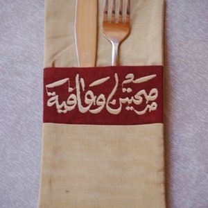 May include: A beige linen napkin with a burgundy band featuring Arabic calligraphy. A silver fork and knife are placed inside the napkin.