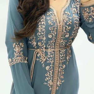 May include: A teal blue kaftan with intricate gold embroidery and beaded trim. The kaftan has a V-neckline and long sleeves.