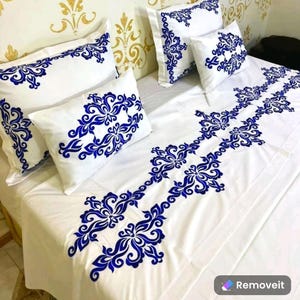 Puede incluir: Ropa de cama blanca con diseños florales bordados en azul. La ropa de cama incluye una sábana plana, una funda de almohada y dos almohadas decorativas.