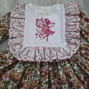 May include: A long-sleeved dress featuring a white bodice with a pink embroidered fairy silhouette holding a heart. The bodice is framed by wide, ruffled lace trim. The skirt and sleeves display a dense floral pattern in shades of brown, pink, and green.