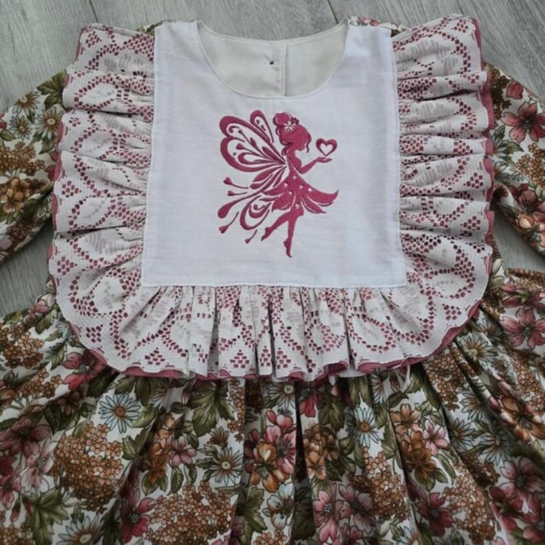 May include: A long-sleeved dress featuring a white bodice with a pink embroidered fairy silhouette holding a heart. The bodice is framed by wide, ruffled lace trim. The skirt and sleeves display a dense floral pattern in shades of brown, pink, and green.