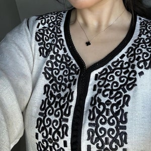 May include: A white linen tunic with black embroidered details. The tunic has a V-neckline and a long sleeve. A black necklace with a clover pendant is worn over the tunic.
