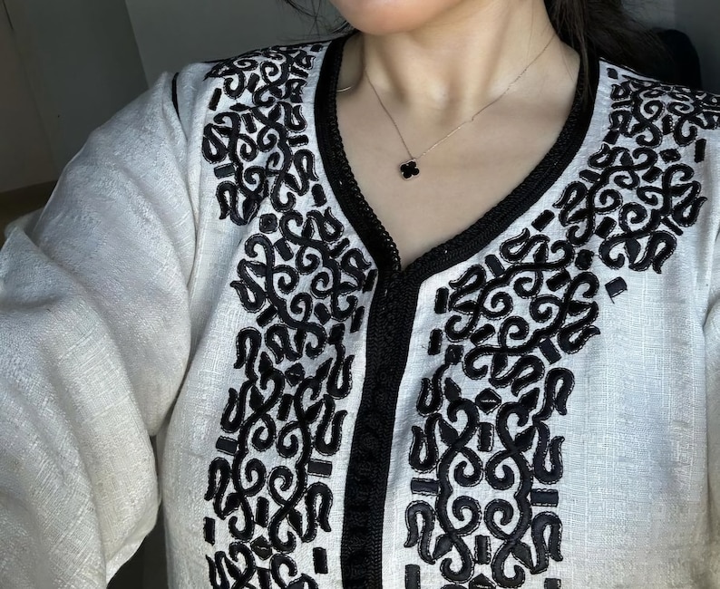 May include: A white linen tunic with black embroidered details. The tunic has a V-neckline and a long sleeve. A black necklace with a clover pendant is worn over the tunic.