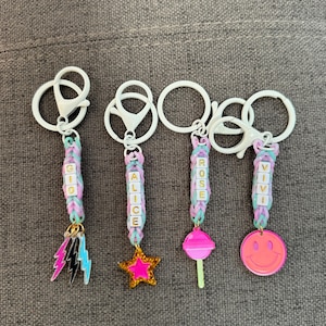 May include: Four colorful keychains with different charms. Each keychain has a name spelled out in colorful beads:  "GIGI", "ALICE", "ROSE", and "VIVI". The charms are a lightning bolt, a star, a lollipop, and a smiley face.