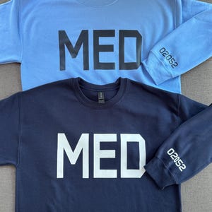 May include: Two crewneck sweatshirts, one light blue and one navy blue. Each features the word "MED" in bold letters across the chest. The light blue sweatshirt has the number "02052" printed on the left sleeve.
