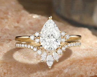 May include: A gold engagement ring set featuring a pear-shaped diamond center stone, surrounded by smaller round and marquise-cut diamonds. The band is also adorned with small diamonds, creating a sparkling effect. The ring is set on a light brown surface.