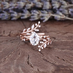 May include: A rose gold engagement ring featuring a large oval-shaped centre stone and a delicate leaf design. The ring is set with smaller diamonds on the band and leaves.
