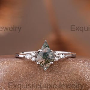 Könnte beinhalten: Ein silberner Ring mit einem rautenförmigen Moosachat-Edelstein, flankiert von kleineren, birnenförmigen Steinen. Der Moosachat weist grüne und braune Einschlüsse auf. Der Ring ist auf einer strukturierten, braunen Oberfläche platziert.