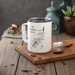 May include: White travel mug with a black outline map of the Florida Keys. The mug features the text "The Florida Keys" and lists the names of the various keys.