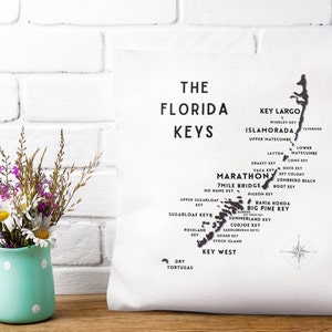 May include: A white throw pillow with a black and white map of the Florida Keys. The map shows the names of the islands and the surrounding waters. The text "The Florida Keys" is printed in black at the top of the pillow.