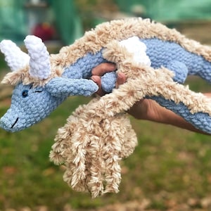 Pode incluir: Um brinquedo de dragão de pelúcia feito à mão em tons de azul e bege. O dragão apresenta um corpo azul com detalhes de pele sintética bege ao longo das costas e da cabeça. Possui chifres brancos e um rosto sorridente, ideal para um brinquedo infantil ou decoração de berçário.
