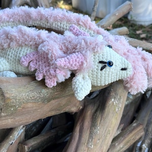 Pode incluir: Um dragão de crochê feito à mão em creme e rosa. O dragão tem um corpo creme, detalhes rosa fofos e olhos azuis. Está deitado em uma pilha de toras de madeira.