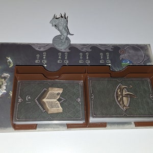 Puede incluir: Un conjunto de componentes de juego de mesa con un tablero con un mapa, tarjeteros marrones y una figura en miniatura. Las cartas representan un libro y un escudo. El tablero tiene un mapa de islas y varios marcadores de juego.