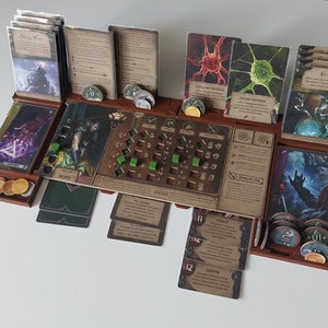Puede incluir: Configuración de un juego de mesa con varias cartas, fichas y un tablero central. Las cartas presentan ilustraciones y texto, mientras que las fichas son de diversas formas y colores. El tablero es marrón.