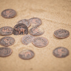 May include: A collection of copper-colored coins with various designs, including elephants and snakes, scattered on a sandy surface.