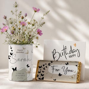 May include: A cylindrical metal tin featuring a butterfly pattern and the text Happy Birthday Sarah A little bit of beautiful in a tin, filled with pink and white wildflowers, alongside a chocolate bar with a butterfly print and Happy Birthday For You text.