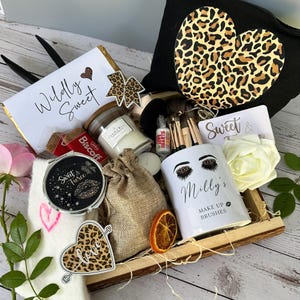 May include: A gift set containing beauty items and sweets. Features a white mug with "Milly's Make Up Brushes" text, a leopard print heart-shaped bag, a candle, and a chocolate bar with "Wildly Sweet" text.