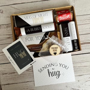 May include: A gift box containing various treats and a card. The box includes a mug, chocolate, a biscuit, and lip balm. The card says "Sending you a hug". A wooden pocket hug is also present.