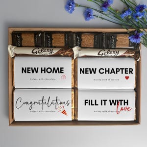 May include: A gift box containing chocolate bars and biscuits. The chocolate bars are printed with messages such as "NEW HOME" and "NEW CHAPTER". Blue flowers decorate the box.