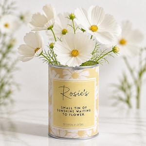 May include: A decorative tin can featuring a yellow label and a daisy pattern, filled with white cosmos flowers. The label says "Rosie's" and "Small tin of sunshine waiting to flower."
