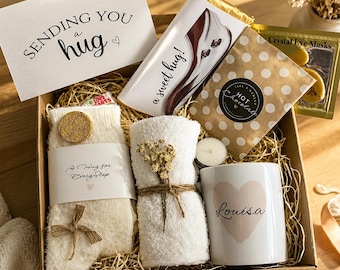 May include: A gift box containing a card reading SENDING YOU a hug, a chocolate bar labeled a sweet hug, a polka dot hot chocolate packet, crystal eye masks, white fuzzy socks with a tag reading A Hug for Every Step, a white towel, and a mug with a heart and the name Louisa.
