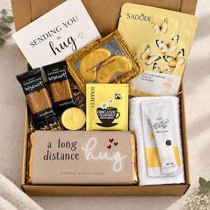 May include: A gift box containing a card reading Sending You a Hug, two caramel biscuits, a gold eye mask, a chamomile tea bag, a yellow candle, a Galaxy milk chocolate bar labeled A Long Distance Hug, a hand cream packet, and a white towel.