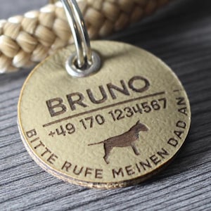 Op de afbeelding: Een tan lederen hondenpenning met de naam "BRUNO" erop gegraveerd. De penning bevat ook een telefoonnummer, " +49 170 1234567", en de tekst "Bitte rufe meinen Daddan!" in het Duits. Een silhouet van een hond is ook gegraveerd op de penning.