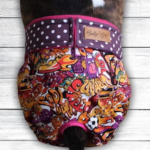 May include: A dog belly band featuring a vibrant graffiti print with cars, flames, and cartoon characters. The top section is dark purple with white polka dots and a brown leather patch labeled Bully Girl. The garment has a pink trim.