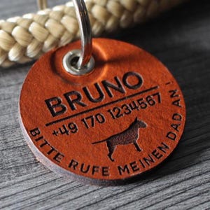 Puede incluir: Una placa de identificación redonda de cuero color canela para mascotas con el nombre BRUNO grabado, el número de teléfono +49 170 1234567, la silueta de un perro y el texto en alemán BITTE RUFE MEINEN DAD AN!, unida a una argolla de metal.