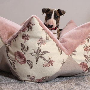 Könnte beinhalten: Ein weißes und rosa Hundebett mit Blumenmuster und einem rosa Kunstfellbesatz. Ein brauner und weißer Bullterrier-Welpe schaut aus dem Bett.