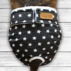 Puede incluir: Pañal para perros negro con estampado de estrellas blancas. El pañal tiene un ribete blanco y una etiqueta de cuero marrón que dice "Bully Girl". Se asegura con cierres de velcro.