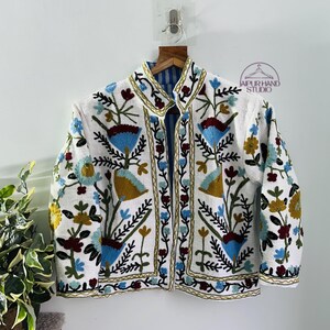 Puede incluir: Chaqueta blanca bordada con un patr&oacute;n floral en tonos de azul, rojo, verde y amarillo. La chaqueta tiene un cuello mandar&iacute;n y cierre de bot&oacute;n. El texto "JAIPUR HAND STUDIO" es visible en la esquina superior derecha de la imagen.