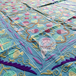 Puede incluir: Un mantel de mesa de algodón azul con bordados intrincados en tonos de rosa, verde y amarillo. El bordado presenta motivos florales y geométricos. El texto "JAIPUR HAND STUDIO" es visible en el centro del mantel.