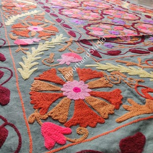 May include: A detailed close-up of a textile with intricate floral embroidery. The design features large, stylized flowers in shades of orange, pink, and red, set against a muted teal background. The embroidery work is visible, showcasing the texture and craftsmanship.