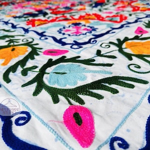 Puede incluir: Primer plano de un textil blanco con bordados coloridos. El diseño presenta patrones florales y geométricos en tonos azules, verdes, amarillos, rosas y naranjas. El bordado es detallado e intrincado, creando una superficie vibrante y texturizada.
