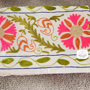 May include: White cotton fabric with embroidered floral designs in pink, orange, and green. The fabric has a blue and white striped border. The text "JAIPUR HAND STUDIO" is embroidered in the bottom right corner.