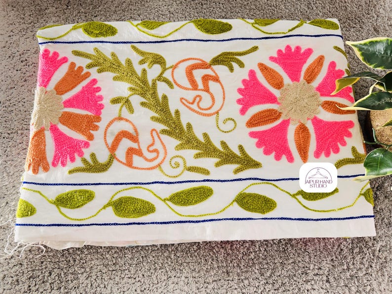 May include: White cotton fabric with embroidered floral designs in pink, orange, and green. The fabric has a blue and white striped border. The text "JAIPUR HAND STUDIO" is embroidered in the bottom right corner.